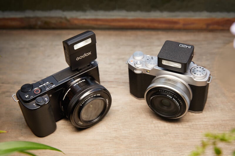 Two compact digital cameras with detachable flashes are placed side by side on a wooden surface. Lenses are attached to both cameras, one in black and the other in silver. The scene is well lit and highlights the camera details.