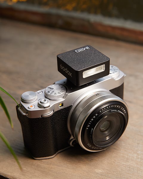 A black and silver vintage style camera with a Godox flash mounted on it, resting on a wooden surface. The camera features a large lens and traditional control dials, with a small plant visible on the side.