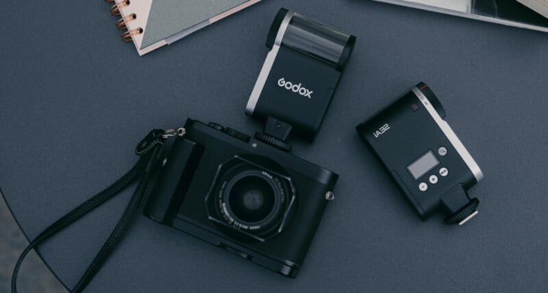 A flat lay of a black camera with a strap next to a Godox flash and small camera accessories on a dark surface. Part of a spiral-bound notebook can be seen in the background.