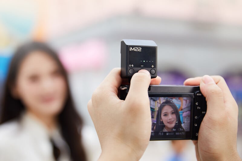 Close-up of hands holding a camera pointed at a person with a clear image of them displayed on the camera screen. The person in the background is out of focus, indicating a shallow depth of field.