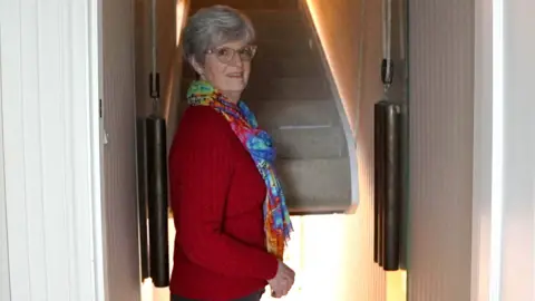 Anthony Leary-White Judith stands at her counterbalanced staircase. The stairs are partially raised and covered with a cream carpet and two weights on the left and right. She is wearing a red sweater, a colorful scarf and dark pants. 