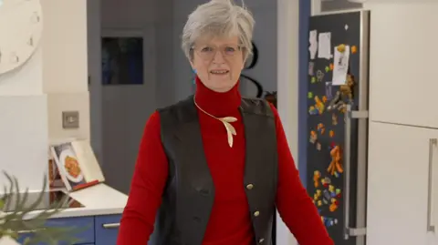 Anthony Leary-White Judith Leary-Joyce stands in her kitchen, looks directly into the camera and smiles. She has short gray hair and wears glasses, a red sweater, a gold leaf necklace and a brown jacket. Behind her is a refrigerator full of magnets and in the left corner lies an open cookbook.