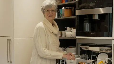 Anthony Leary-White Judith Leary-Joyce unloads her dishwasher and smiles into the camera. She has white hair and glasses and is wearing a long white knit sweater.