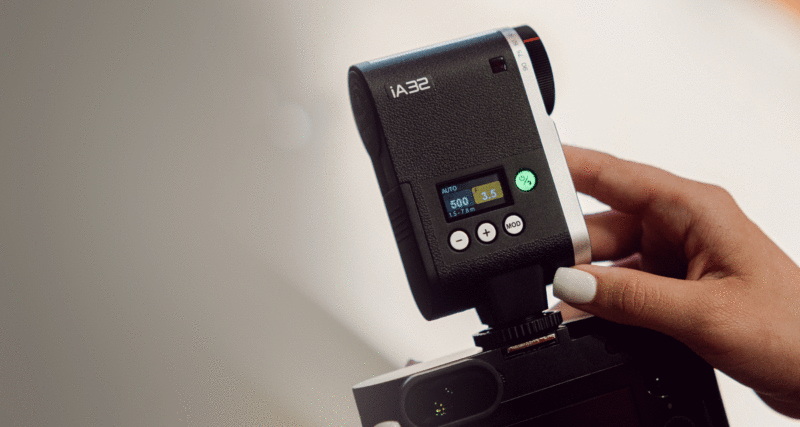 A person with painted nails adjusts a mounted iA32 camera light with a digital display showing the settings. The light is attached to a camera and stands against a soft focus background.