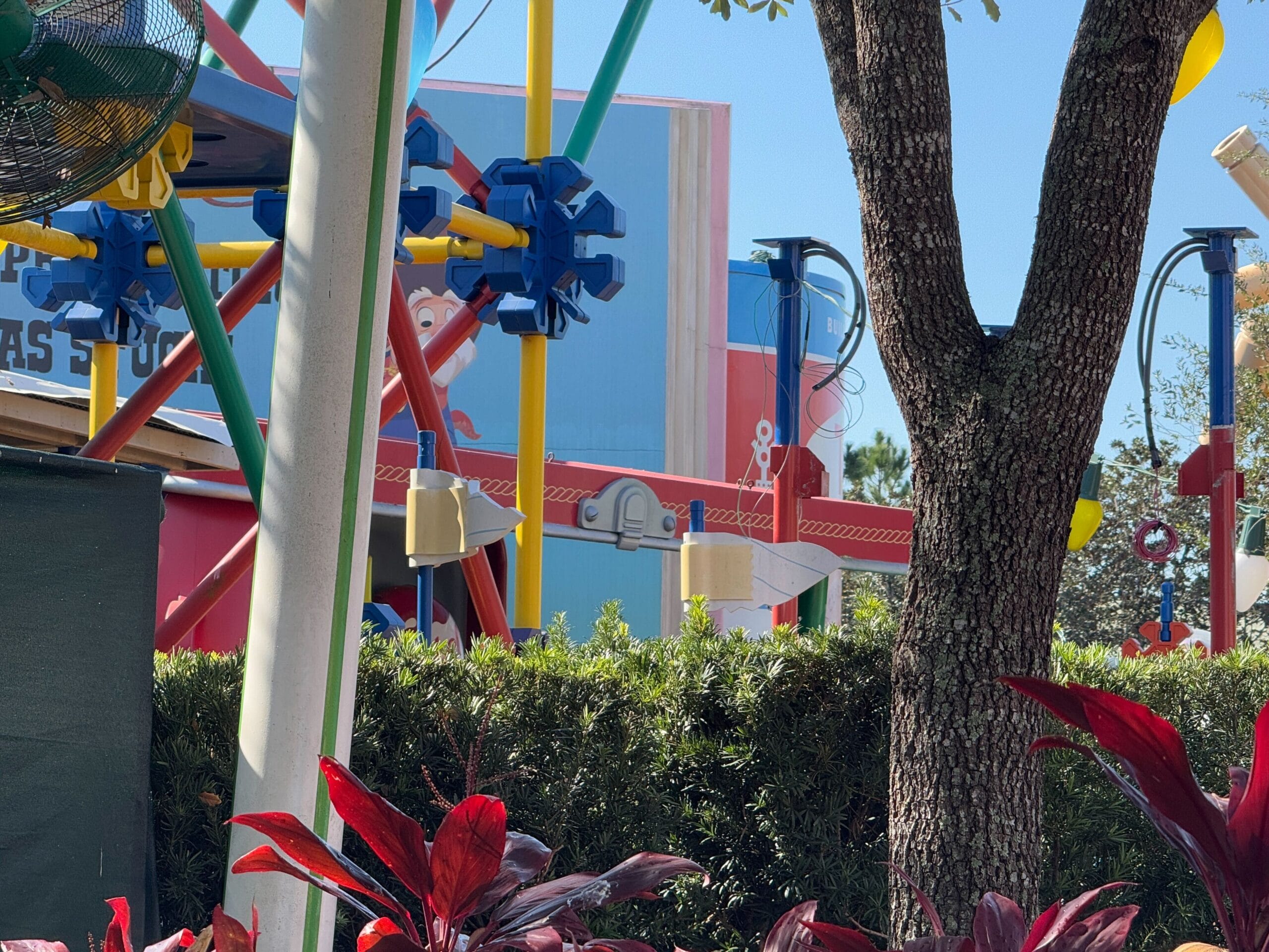 Amusement parking with colorful structures and gears, surrounded by lush green and red-leafed plants, which offer a perfect view from the temporary seating area near Woody's lunch box.