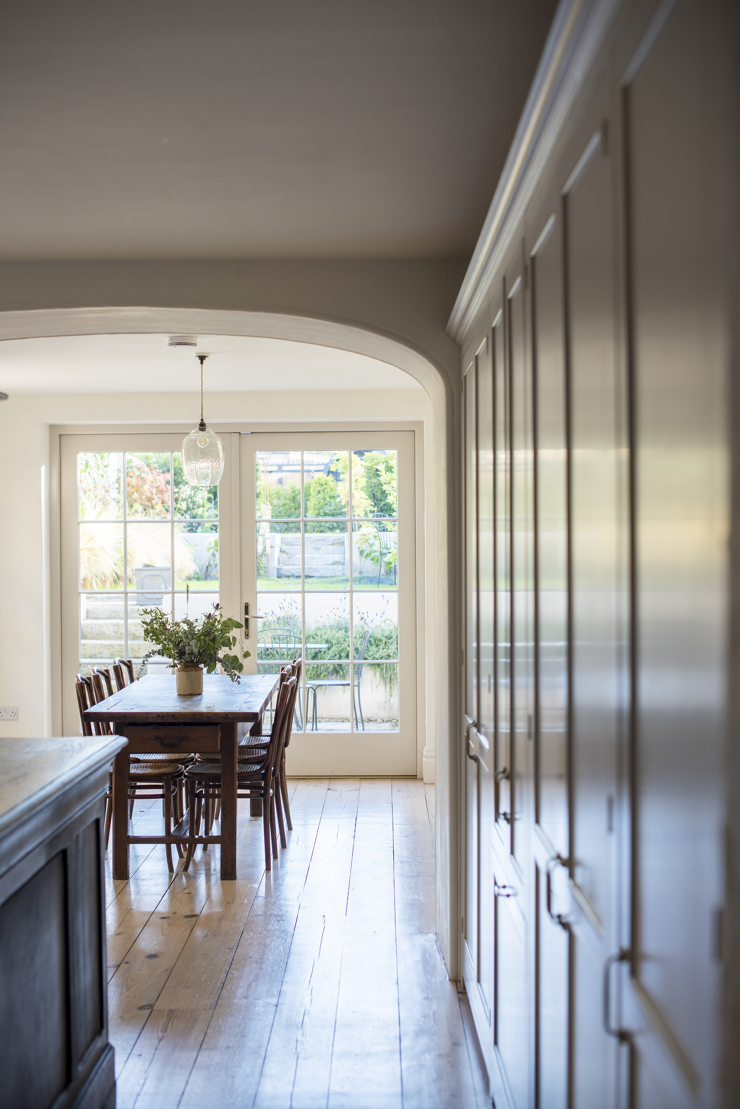 Wooden dining table with chairs in a light room, observed through a arched door.