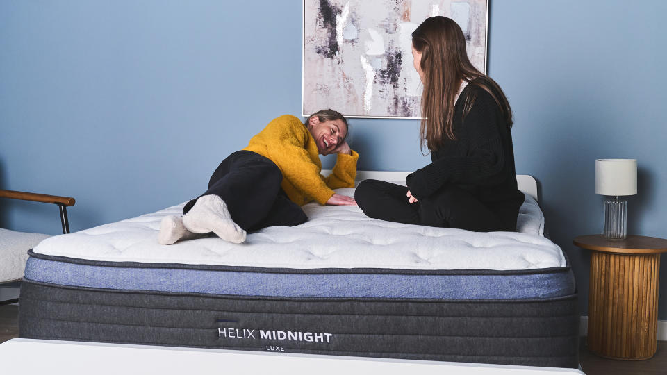 A few product testers over the Helix Midnight Luxe mattress, with one person lying on their side and the other person sits next to him