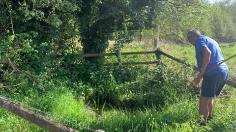 Helen Heywood A man with a blue t -shirt and shorts that hold a long stick and stand in the long grass with a wooden fence, surrounds it