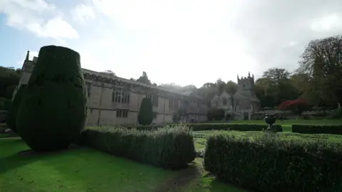 BBC Lanhydrock with bushes surrounding the building, there is a castle building with turfen on the right. On the right there is a large hedge that was shaped.