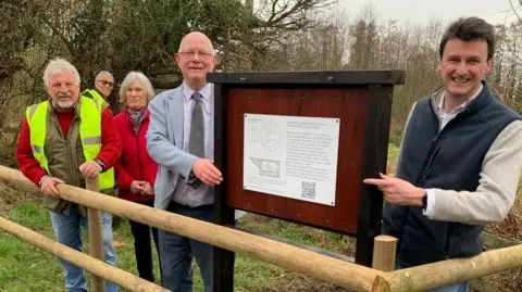 Helen Heywood Four men and one woman stood in front of some trees behind a wooden fence next to a wooden shield with a white badge on it