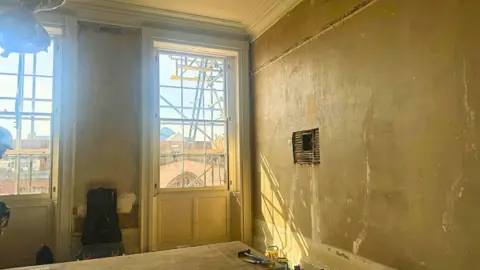 BBC/ Emily Johnson a room with bare walls and floor to ceiling windows. The scaffold can be seen through the windows.