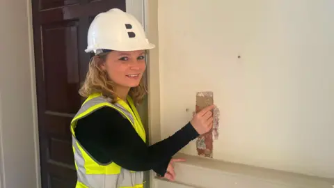 BBC/ Emily Johnson, a woman, refers to exposed layers of paint on a white wall in a Hi-vis vest and white helmet.