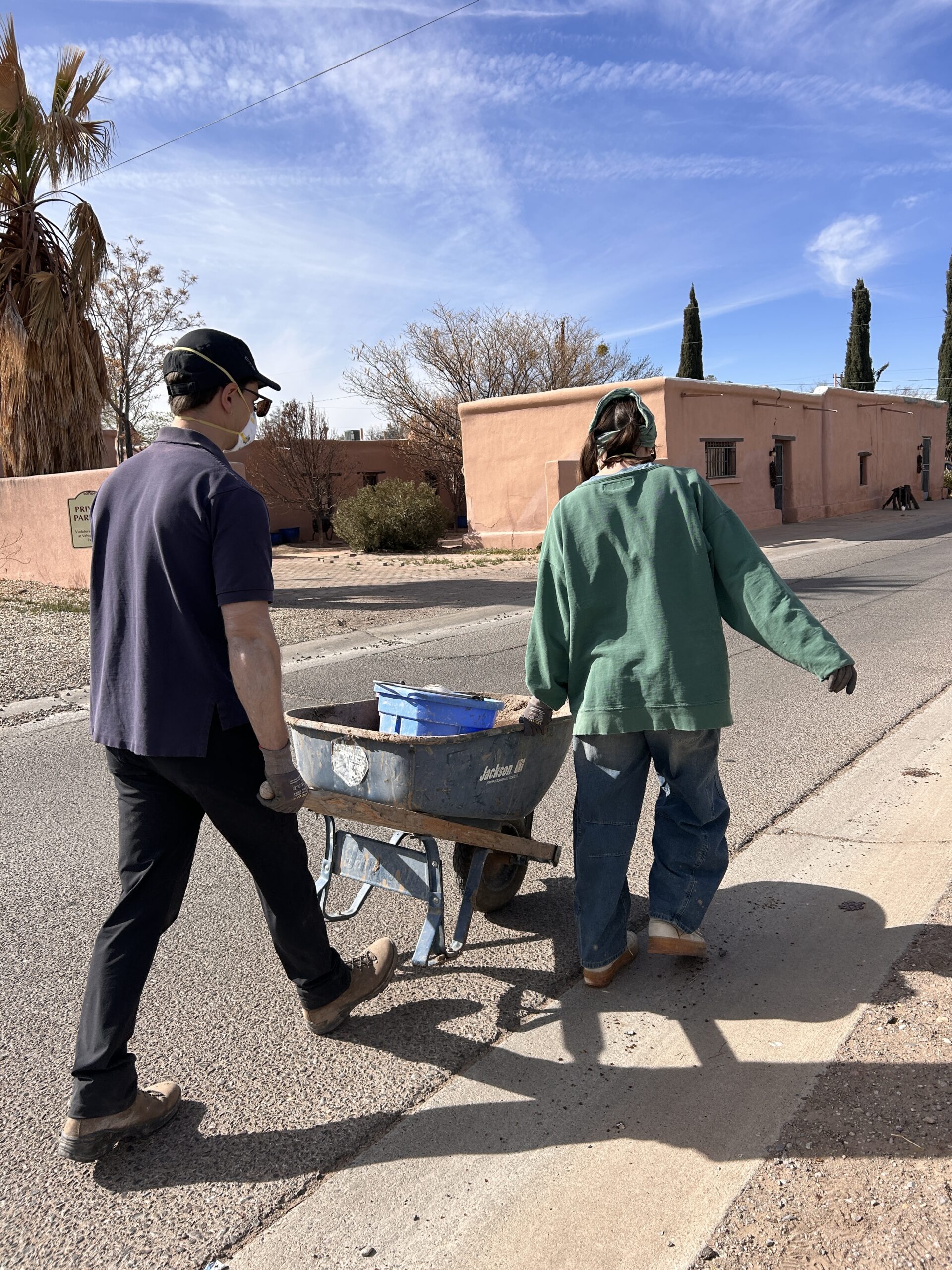 A person who wears a yellow baseball cap, safety glasses, a mask, a blue plaid jacket, pink trousers and brown boots uses a power drilling machine to mix a substance in a bucket. They work in an earthly -walled building with two other people in the background who also wear masks and hats.