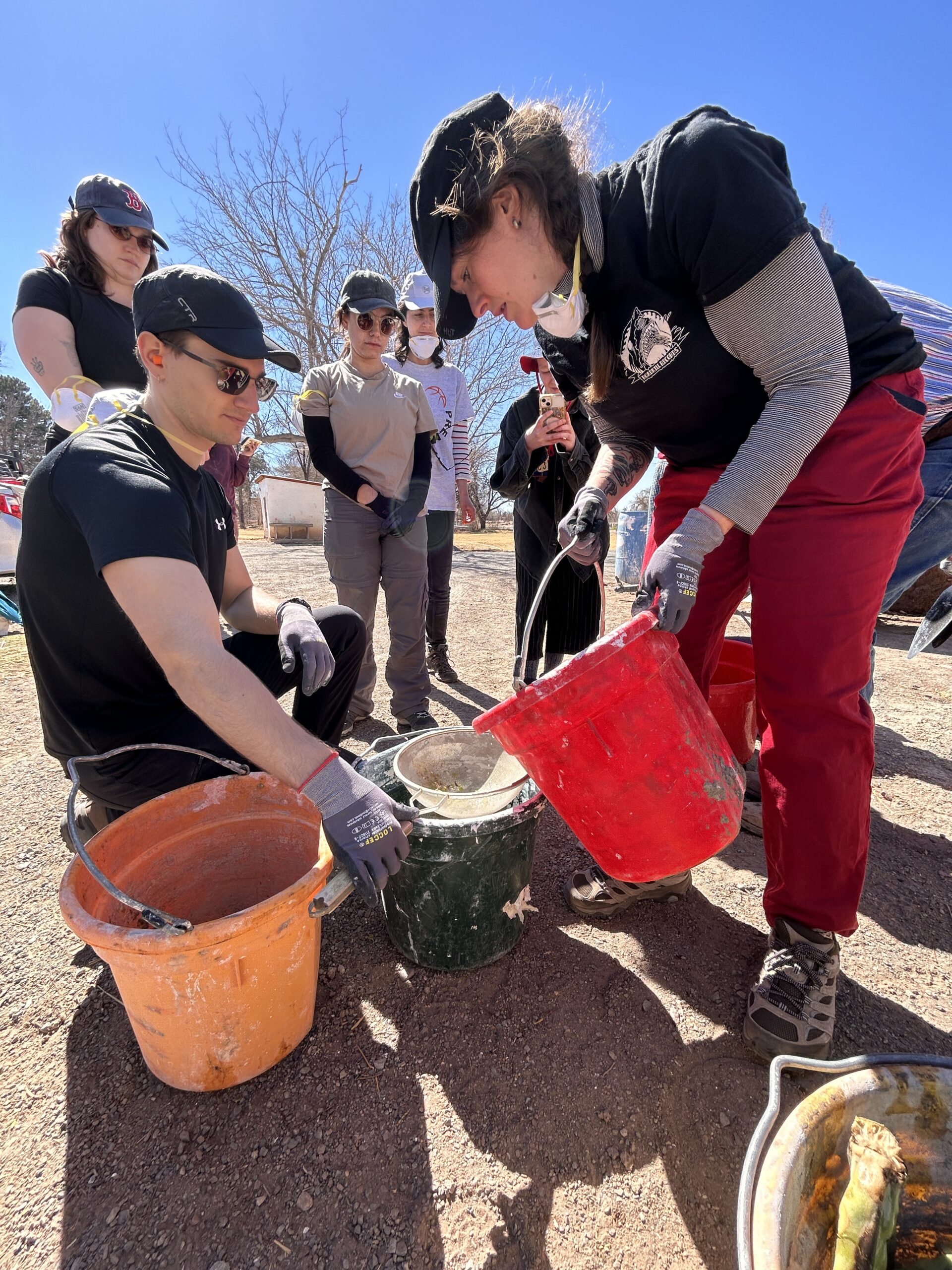 A group of people outdoors under a clear blue sky works with buckets and building materials. A woman in a black hat, a black shirt and red trousers pour a red bucket into a green bucket, while a man who wears a black hat and sunglasses that keeps the bucket calm. Some others who wear gloves, masks and casual work clothes observe the process.