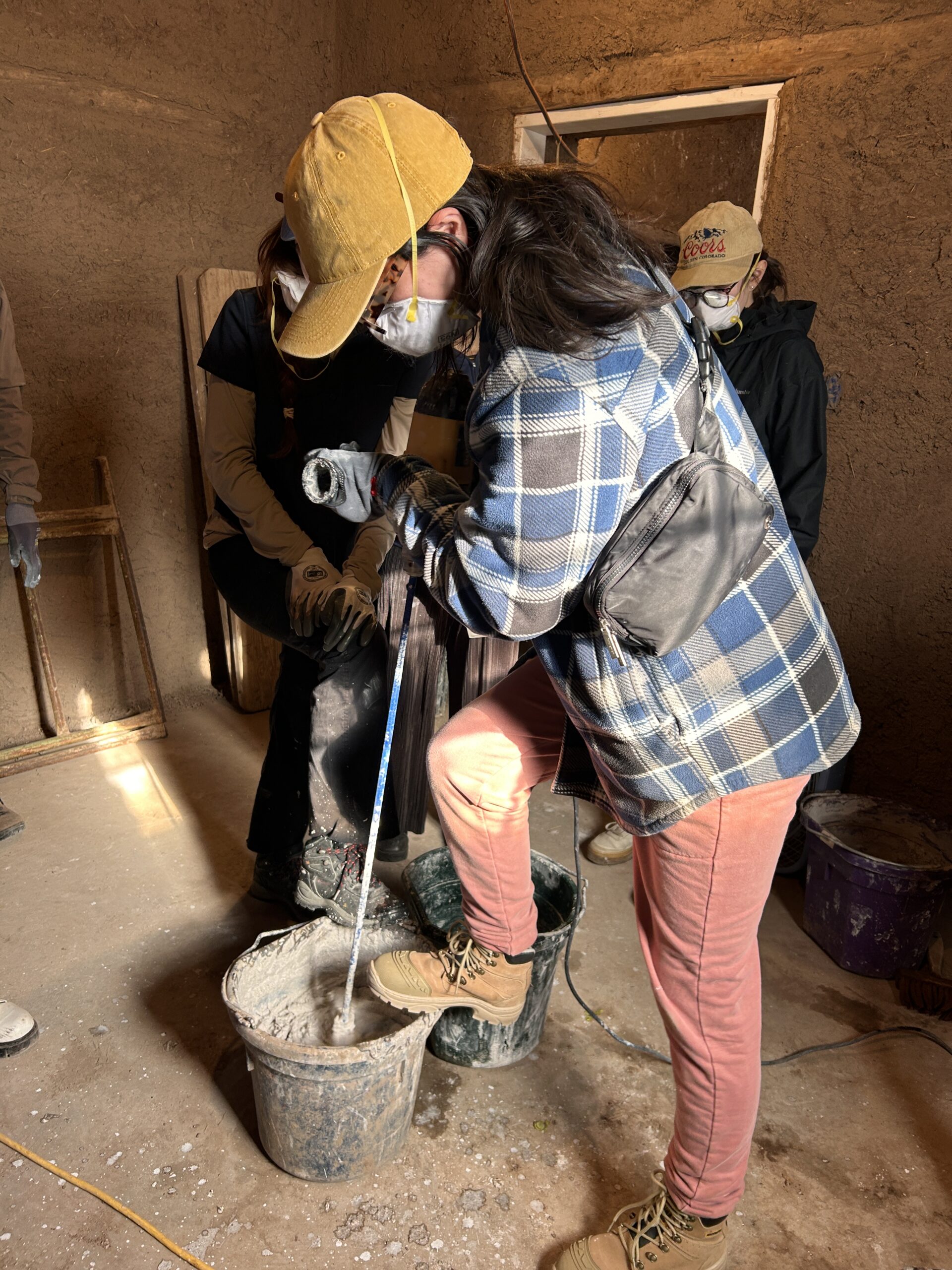 A person who wears a yellow baseball cap, safety glasses, a mask, a blue plaid jacket, pink trousers and brown boots uses a power drilling machine to mix a substance in a bucket. They work in an earthly -walled building with two other people in the background who also wear masks and hats.