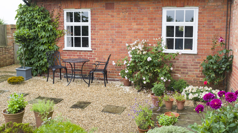 A brick house has a gravel terrace surrounded by blooming plants
