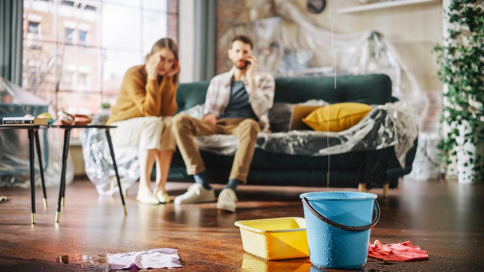 The roof is leaking at home or pipe rupture: couple in despair on a sofa and observes how water drips into buckets in your living room. Disaster, distal and financial ruin