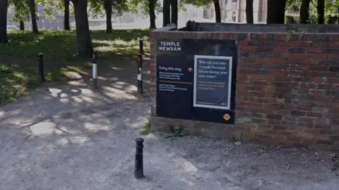 Google a sign on a brick wall that specifies the way to the Temple Newsam. Traffic seamies and trees can be seen in the background.
