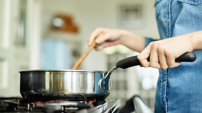 View the middle section below Image of women who cook food in the pan. Utensil is placed on gas stove. Woman stirs the bowl in the pan.