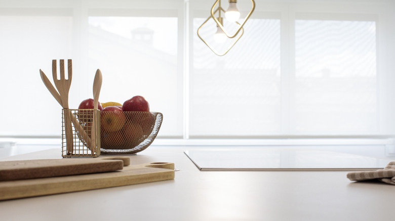 White kitchen counter with bowl of apples, wooden cutting boards and wooden utensils.