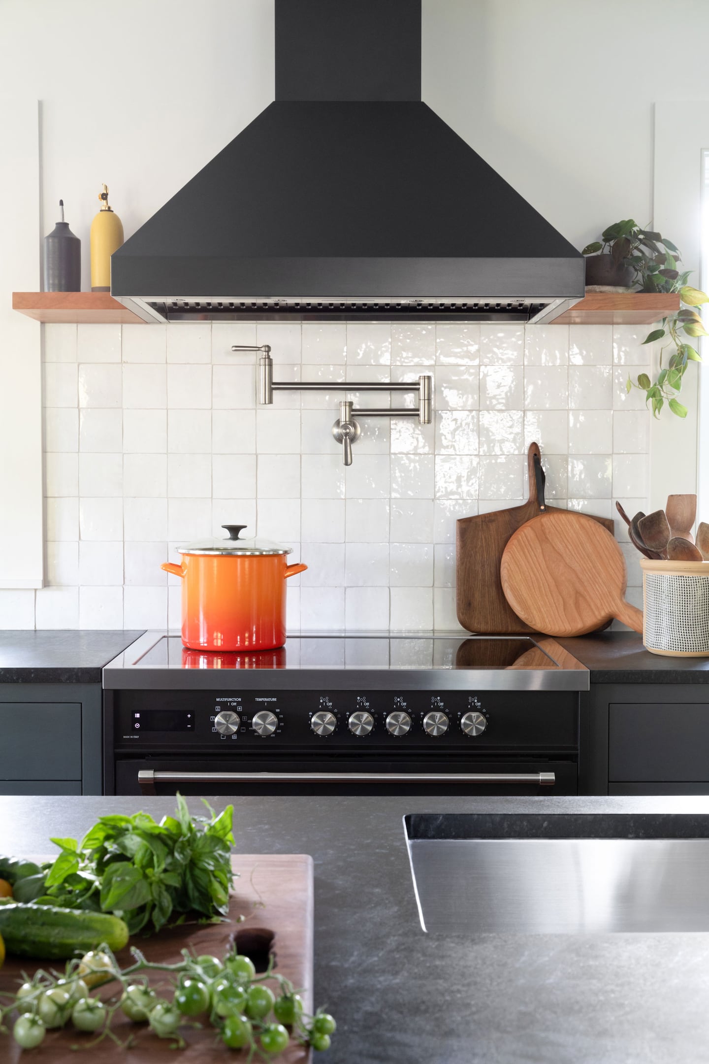 In the background you can see a black stove with an orange pot on the front left stove plate. There is a black extractor hood above the hob with small wooden shelves on either side. On the shelf to the left there are two bottles with spouts for cooking oils and on the shelf to the right there is a plant. The back wall is made of white tiles and there is a tap on the wall for filling large pans. In the foreground is a prep island with a black countertop and sink. There are herbs on a cutting board on the worktop.